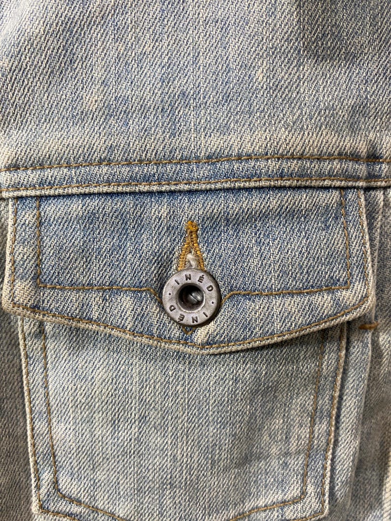 May include: A light blue denim jacket pocket featuring a weathered texture and gold-toned stitching. A circular, dark metallic button is centered on the pocket flap, embossed with the text INED in a circular pattern around the center hole.