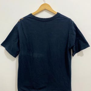 May include: A dark navy blue short-sleeve t-shirt hanging on a wooden hanger. The shirt features a crew neckline and a raw, unfinished hemline at the bottom. A small portion of a yellow and orange graphic design is visible on the upper left shoulder.