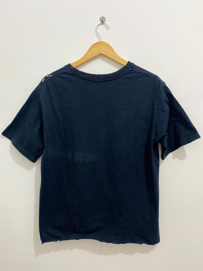 May include: A dark navy blue short-sleeve t-shirt hanging on a wooden hanger. The shirt features a crew neckline and a raw, unfinished hemline at the bottom. A small portion of a yellow and orange graphic design is visible on the upper left shoulder.