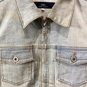 May include: A light-wash blue denim jacket featuring a full-length front zipper, a classic collar, and two chest pockets with button closures. The interior neck label displays the text INED MADE IN JAPAN, and the metal buttons are embossed with text.