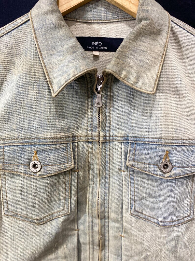 May include: A light-wash blue denim jacket featuring a full-length front zipper, a classic collar, and two chest pockets with button closures. The interior neck label displays the text INED MADE IN JAPAN, and the metal buttons are embossed with text.