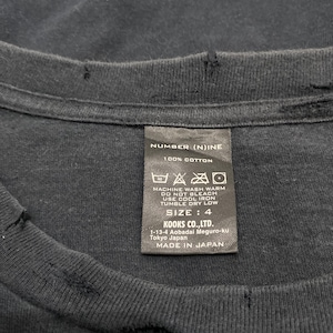 May include: A black cotton t-shirt with a distressed, frayed neckline and a rectangular care label. The label reads NUMBER (N)INE, 100% COTTON, SIZE: 4, KOOKS CO., LTD. 1-13-4 Aobadal Meguro-ku Tokyo Japan, and MADE IN JAPAN with care symbols.