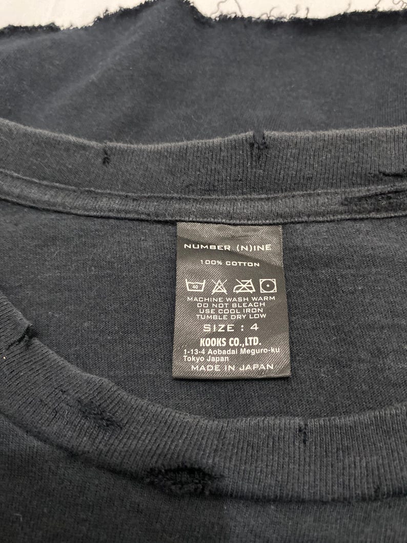 May include: A black cotton t-shirt with a distressed, frayed neckline and a rectangular care label. The label reads NUMBER (N)INE, 100% COTTON, SIZE: 4, KOOKS CO., LTD. 1-13-4 Aobadal Meguro-ku Tokyo Japan, and MADE IN JAPAN with care symbols.