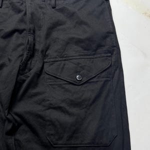 Puede incluir: Primer plano de pantalones negros con un bolsillo abotonado en el lateral. Los pantalones tienen cierre de bot&oacute;n y cremallera en la cintura. La tela parece ser un material tejido resistente. Los pantalones son de color negro s&oacute;lido.