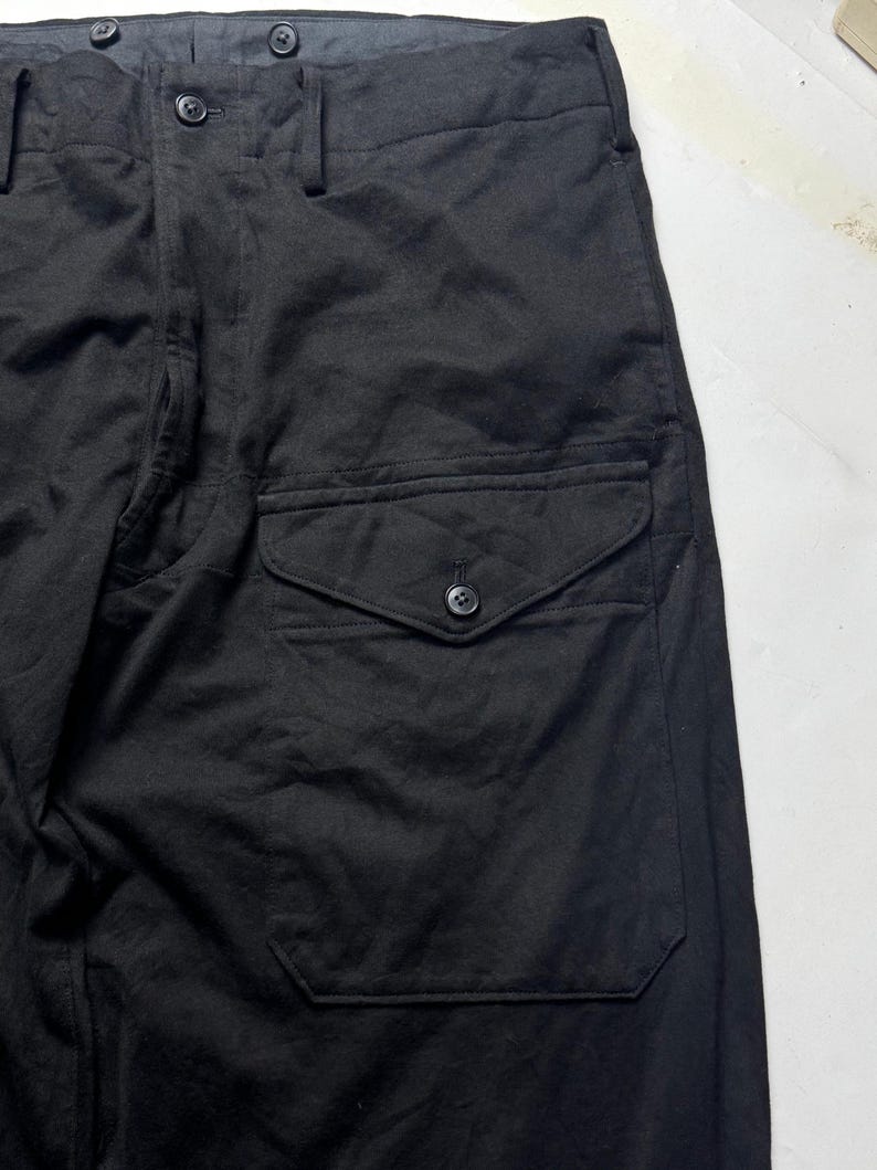 Puede incluir: Primer plano de pantalones negros con un bolsillo abotonado en el lateral. Los pantalones tienen cierre de bot&oacute;n y cremallera en la cintura. La tela parece ser un material tejido resistente. Los pantalones son de color negro s&oacute;lido.