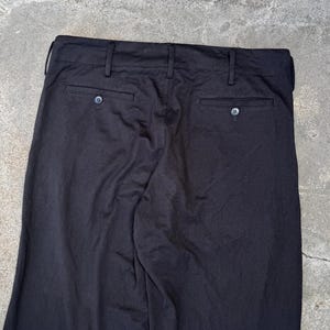 Puede incluir: Pantalones negros con dos bolsillos traseros y cierres de bot&oacute;n. Los pantalones tienen trabillas y un dise&ntilde;o cl&aacute;sico. La tela parece ser un material de peso medio, adecuado para diversas ocasiones. Se ve una cinta m&eacute;trica amarilla.