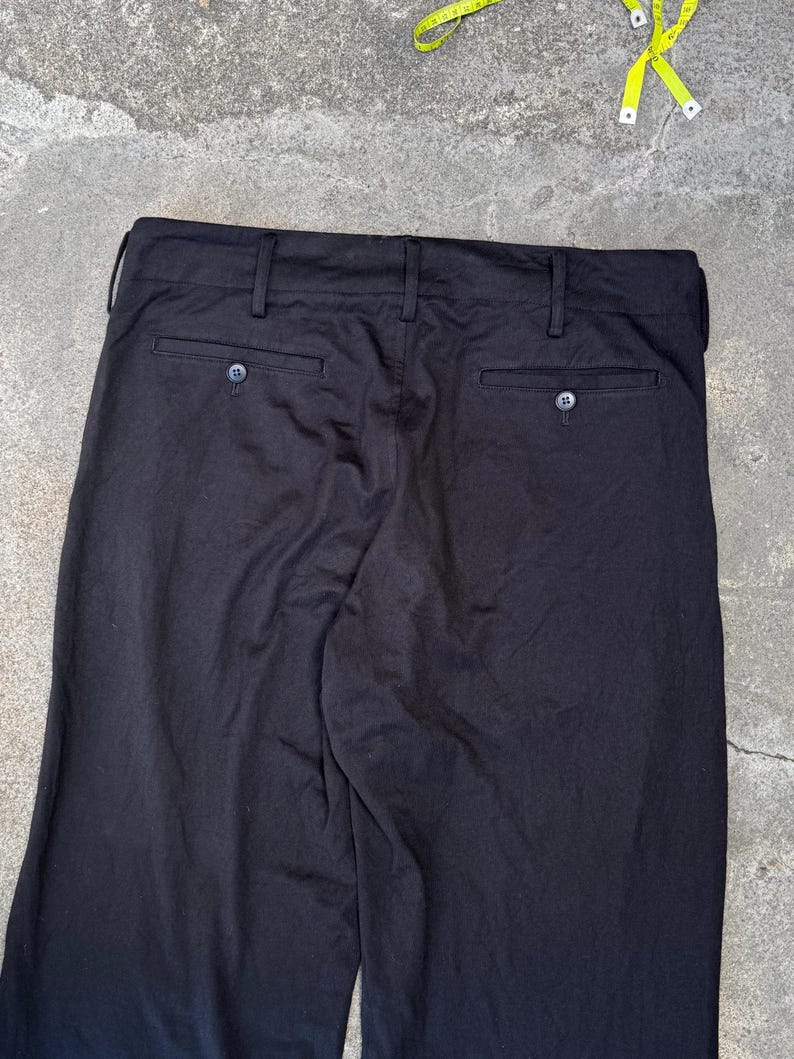 Puede incluir: Pantalones negros con dos bolsillos traseros y cierres de bot&oacute;n. Los pantalones tienen trabillas y un dise&ntilde;o cl&aacute;sico. La tela parece ser un material de peso medio, adecuado para diversas ocasiones. Se ve una cinta m&eacute;trica amarilla.