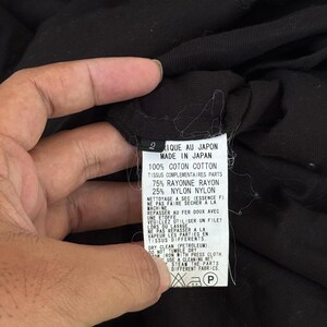 May include: A black garment tag with text in French and English, including "MADE IN JAPAN" and fabric composition details. The tag is held by a hand, with the garment's fabric visible in the background.