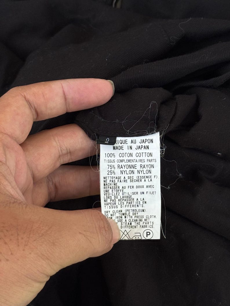 May include: A black garment tag with text in French and English, including "MADE IN JAPAN" and fabric composition details. The tag is held by a hand, with the garment's fabric visible in the background.