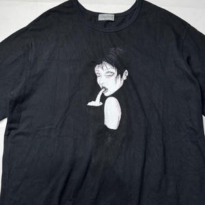 May include: Black t-shirt featuring a stylized illustration of a person with dark hair and pale skin. The graphic is in black and white, with a dark background. The shirt has a crew neck and short sleeves.