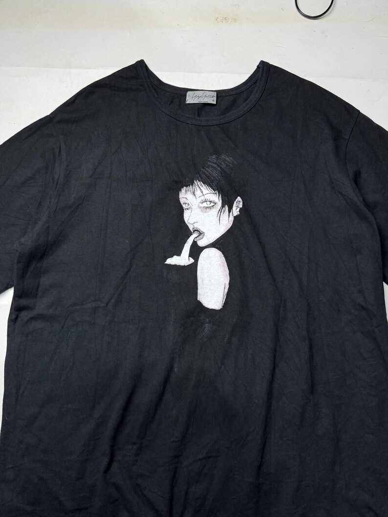 May include: Black t-shirt featuring a stylized illustration of a person with dark hair and pale skin. The graphic is in black and white, with a dark background. The shirt has a crew neck and short sleeves.