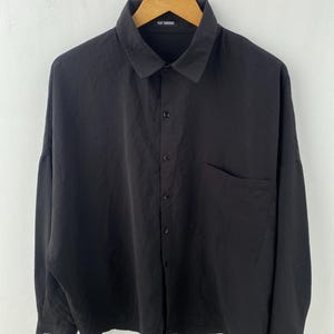 May include: Black button-down shirt with a single chest pocket. The shirt has long sleeves and a classic collar. The brand name "RAF SIMONS" is visible on the collar.