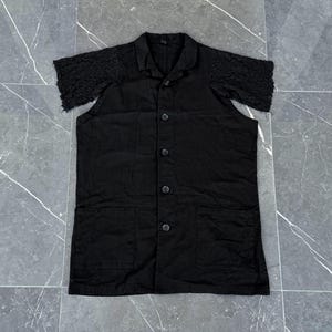May include: A black short-sleeved button-down shirt with a collar and two front pockets. The sleeves are made of a textured, lace-like fabric. The shirt has a classic design with a modern twist.