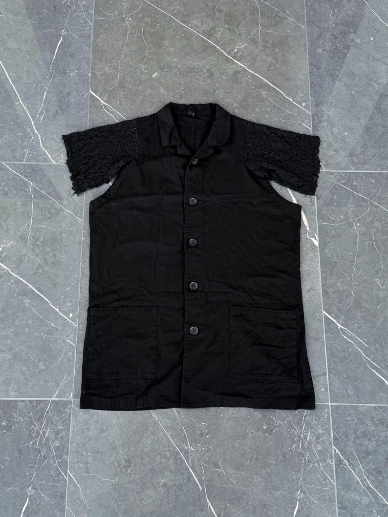 May include: A black short-sleeved button-down shirt with a collar and two front pockets. The sleeves are made of a textured, lace-like fabric. The shirt has a classic design with a modern twist.