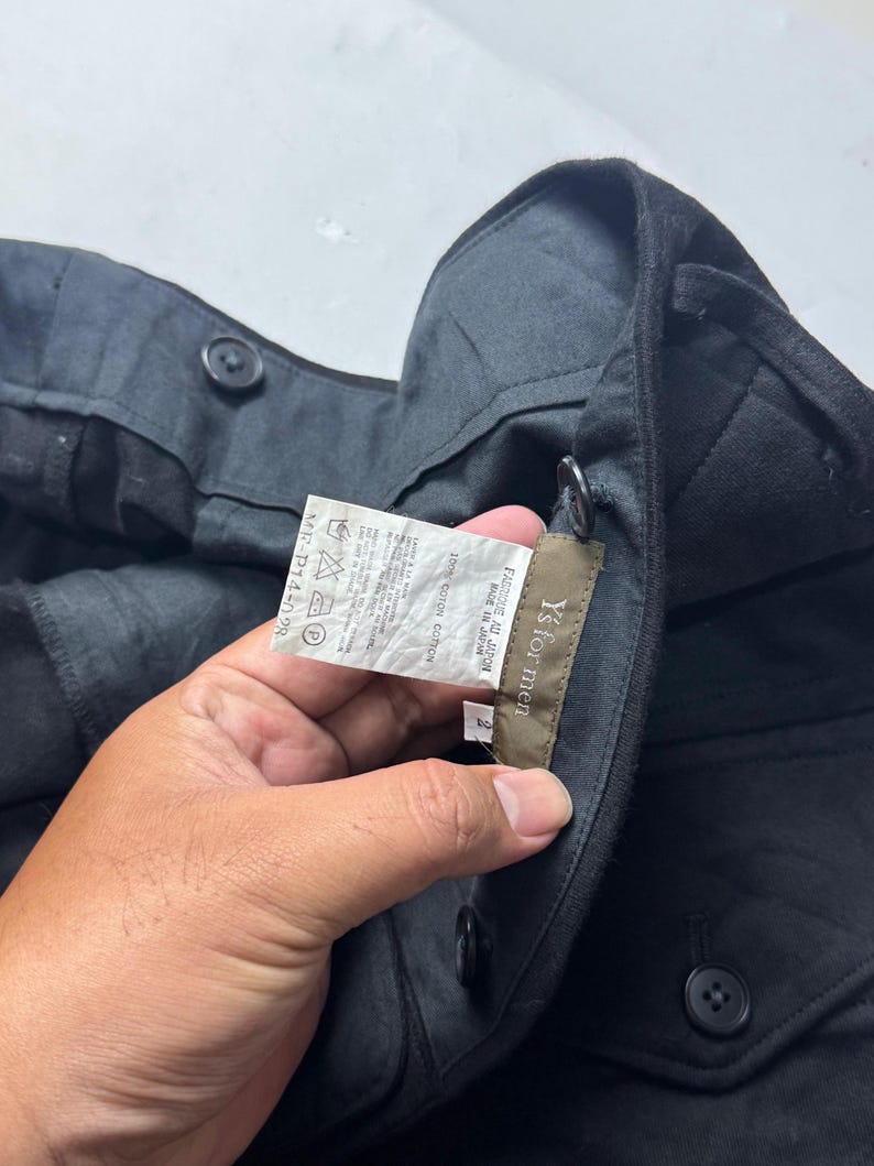 Puede incluir: Pantalones negros con botones. La etiqueta dice "Y's for men", "FABRIQUE AU JAPON" y "MADE IN JAPAN". La tela es 100% algod&oacute;n. Los pantalones tienen una etiqueta con instrucciones de cuidado y el c&oacute;digo MF-P14-028.