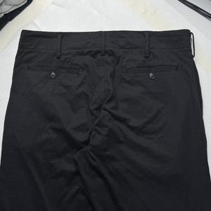 Puede incluir: Pantalones negros con dos bolsillos traseros y cierres de bot&oacute;n. Los pantalones tienen trabillas y un dise&ntilde;o cl&aacute;sico. La tela parece ser un material de peso medio, adecuado para diversas ocasiones.