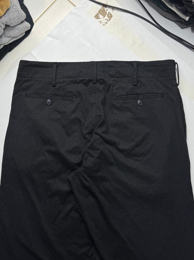 Puede incluir: Pantalones negros con dos bolsillos traseros y cierres de bot&oacute;n. Los pantalones tienen trabillas y un dise&ntilde;o cl&aacute;sico. La tela parece ser un material de peso medio, adecuado para diversas ocasiones.