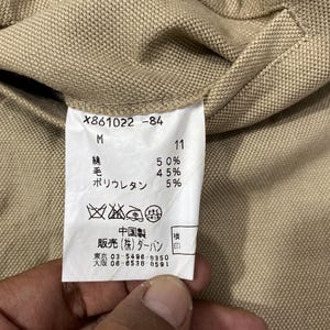 Puede incluir: Primer plano de una tela beige con una etiqueta blanca. La etiqueta contiene texto como "X861022-84", "M" y porcentajes de algod&oacute;n, lana y poliuretano. Tambi&eacute;n hay caracteres japoneses y s&iacute;mbolos de cuidado.
