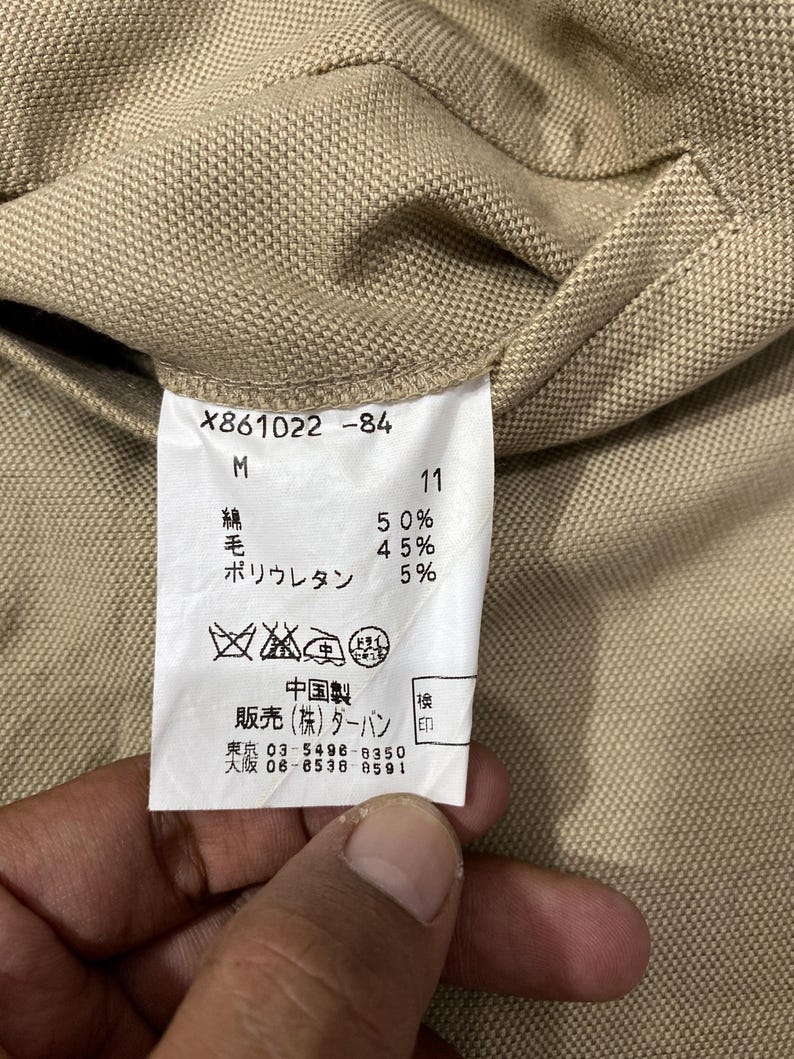 Puede incluir: Primer plano de una tela beige con una etiqueta blanca. La etiqueta contiene texto como "X861022-84", "M" y porcentajes de algod&oacute;n, lana y poliuretano. Tambi&eacute;n hay caracteres japoneses y s&iacute;mbolos de cuidado.