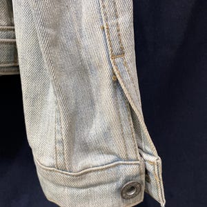 May include: A light-wash blue denim jacket sleeve featuring a structured cuff with a single metallic button. The fabric displays a visible diagonal twill weave pattern with reinforced stitching along the seams and a classic buttoned wrist closure.