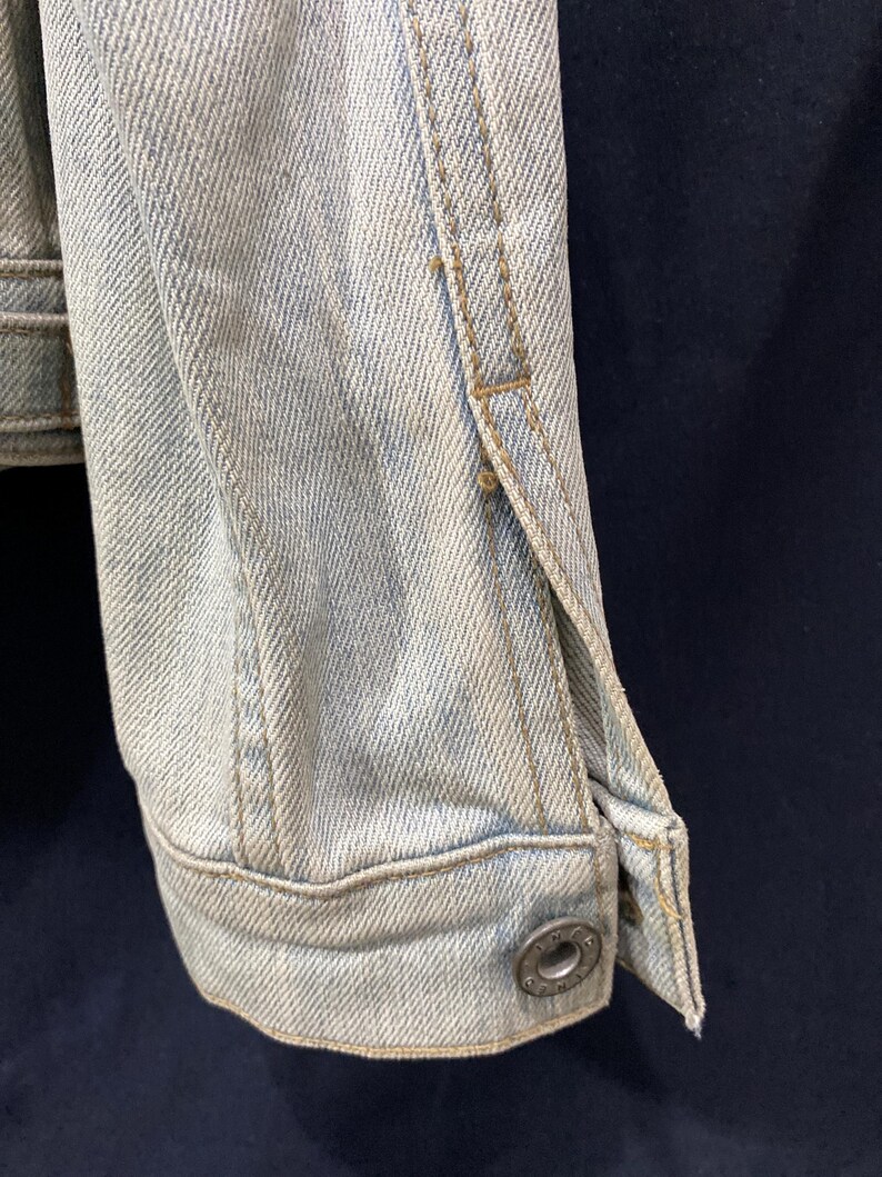 May include: A light-wash blue denim jacket sleeve featuring a structured cuff with a single metallic button. The fabric displays a visible diagonal twill weave pattern with reinforced stitching along the seams and a classic buttoned wrist closure.