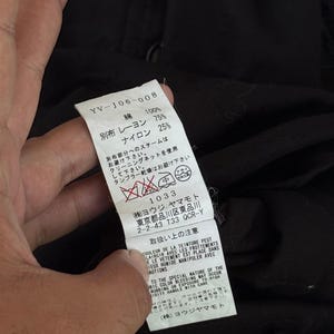 May include: A close-up of a white tag with Japanese text and care instructions. The tag also includes the text "YV-J06-008", fabric composition, and a care symbol. The tag is held against a black fabric background.