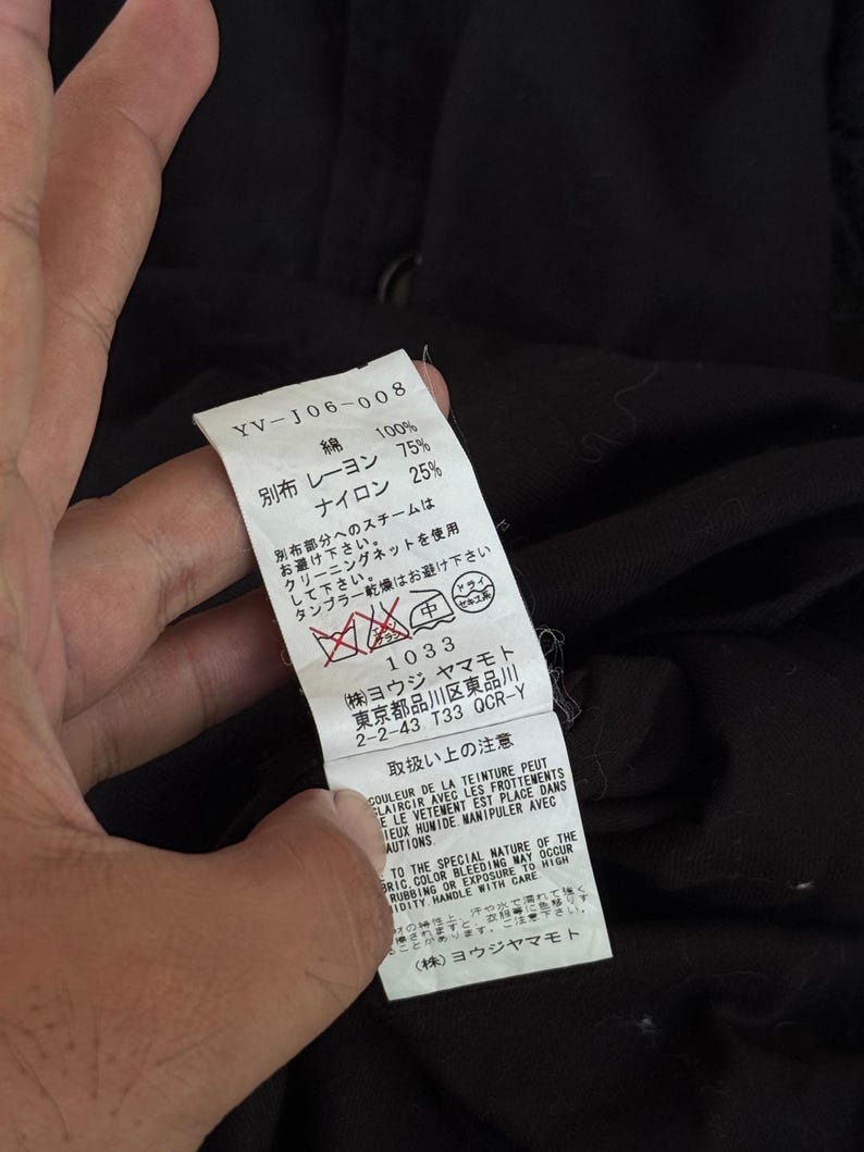 May include: A close-up of a white tag with Japanese text and care instructions. The tag also includes the text "YV-J06-008", fabric composition, and a care symbol. The tag is held against a black fabric background.