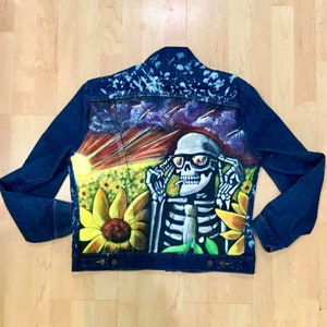 Könnte beinhalten: Eine blaue Jeansjacke mit einem bemalten Design eines Skeletts, das eine Sonnenbrille trägt und eine Zigarette raucht. Das Skelett ist von Sonnenblumen und einem Sonnenuntergangshintergrund umgeben.