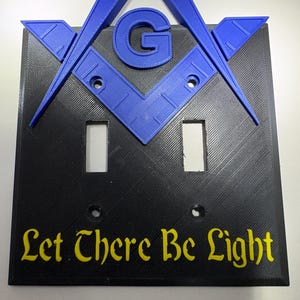 May include: A black light switch cover with a blue Masonic symbol and the text "Let There Be Light" in yellow. The cover has two rectangular cutouts for the switches and two screw holes.