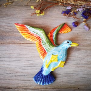 May include: A ceramic wall hanging of a blue bird with yellow, orange and green wings. The bird is in flight with its wings spread out.