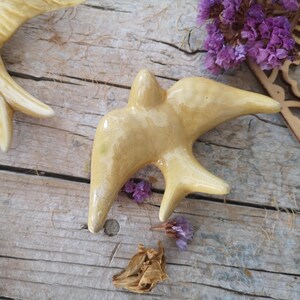 May include: A ceramic bird figurine in a light yellow glaze, with outstretched wings and a detailed form. The bird is set on a weathered wooden surface, with dried flowers and a decorative wooden cutout nearby. The overall theme is rustic decor.