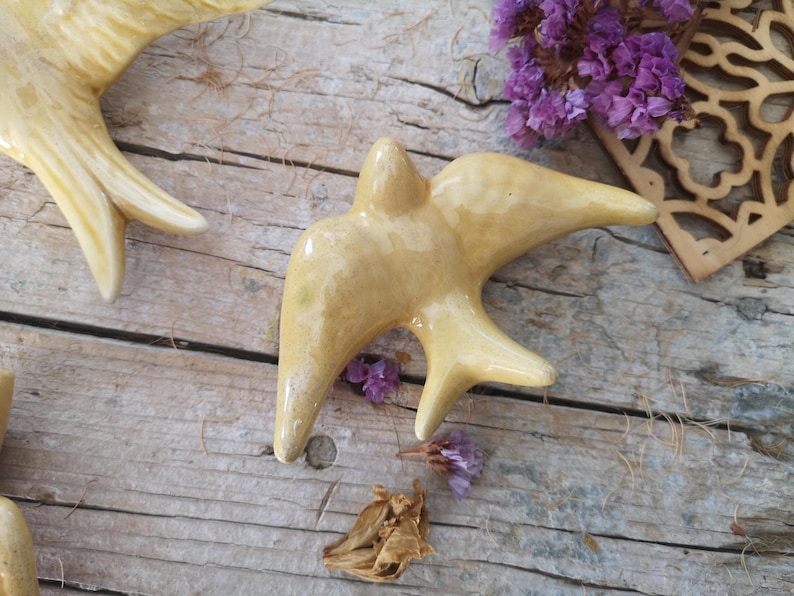 May include: A ceramic bird figurine in a light yellow glaze, with outstretched wings and a detailed form. The bird is set on a weathered wooden surface, with dried flowers and a decorative wooden cutout nearby. The overall theme is rustic decor.