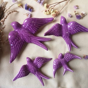 Puede incluir: Cuatro pájaros de cerámica púrpura con alas detalladas y un acabado texturizado. Los pájaros están dispuestos en un patrón decorativo sobre una superficie blanca.