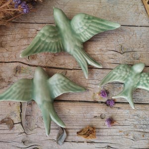 May include: Three ceramic swallow birds in a light green colour, mounted on a wooden surface.
