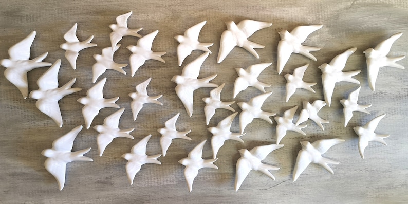 Op de afbeelding: Een verzameling witte keramische zwaluwen in verschillende maten, gerangschikt op een lichtgrijze houten ondergrond. De vogels zijn in vlucht, met gespreide vleugels, waardoor een decoratieve weergave ontstaat. De zwaluwen zijn glad en gedetailleerd.
