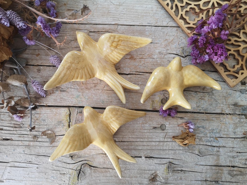May include: Three ceramic swallow figurines in a warm beige color with outstretched wings are arranged on a weathered wooden surface. The birds are detailed with textured wings and tail feathers. Dried flowers and decorative wooden cutouts surround the birds.
