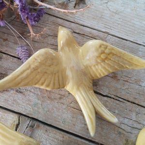 May include: A ceramic swallow figurine with outstretched wings, in a creamy yellow color. The bird is detailed with etched wing feathers and a forked tail. The figurine is set against a rustic wooden background, with dried purple flowers.