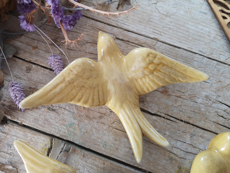 May include: A ceramic swallow figurine with outstretched wings, in a creamy yellow color. The bird is detailed with etched wing feathers and a forked tail. The figurine is set against a rustic wooden background, with dried purple flowers.
