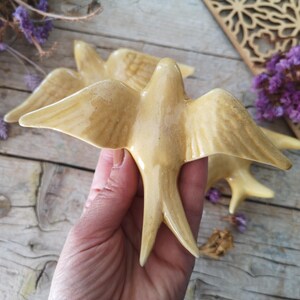 May include: Ceramic swallow figurines in a warm, creamy yellow hue. The birds have outstretched wings and detailed feather patterns. The figurines are held in a hand, with additional birds visible in the background. Dried purple flowers and a wooden decorative piece are also present.