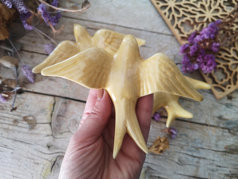 May include: Ceramic swallow figurines in a warm, creamy yellow hue. The birds have outstretched wings and detailed feather patterns. The figurines are held in a hand, with additional birds visible in the background. Dried purple flowers and a wooden decorative piece are also present.