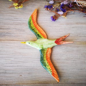 May include: A ceramic bird wall decor with green, orange, and yellow wings. The bird is in flight and has a yellow beak.