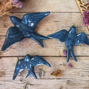 Puede incluir: Tres piezas de decoración de pared de golondrina de cerámica azul. Las aves están en varias poses con las alas extendidas. Las aves son de color azul oscuro con un acabado ligeramente texturizado.