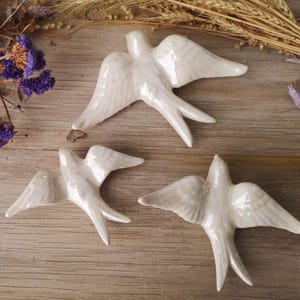 Puede incluir: Tres golondrinas de cerámica blanca con las alas extendidas están dispuestas sobre una superficie de madera. Las aves tienen texturas de plumas detalladas y están colocadas cerca de flores secas y espigas de trigo. Las golondrinas son un objeto decorativo.