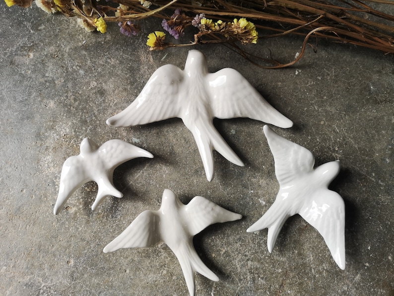 Op de afbeelding: Vier witte keramische zwaluwen met uitgespreide vleugels en gedetailleerde veertjes zijn op een grijze ondergrond geplaatst. De vogels staan in verschillende poses. Gedroogde bloemen zijn op de achtergrond. Decoratieve items.