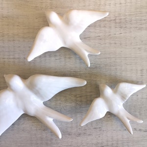Op de afbeelding: Drie witte keramische zwaluwen met uitgespreide vleugels, geplaatst op een licht oppervlak met houtnerf. De vogels zijn gedetailleerd en elegant. Een decoratief item voor in huis, dat een gevoel van vlucht en vrijheid oproept.