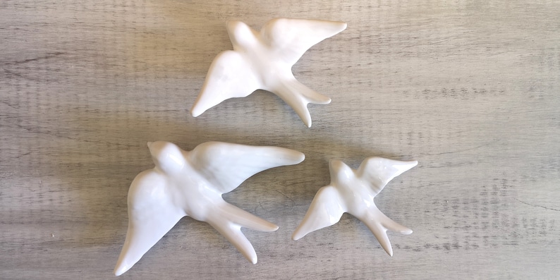 Op de afbeelding: Drie witte keramische zwaluwen met uitgespreide vleugels, geplaatst op een licht oppervlak met houtnerf. De vogels zijn gedetailleerd en elegant. Een decoratief item voor in huis, dat een gevoel van vlucht en vrijheid oproept.