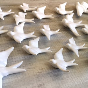 Op de afbeelding: Een verzameling witte keramische vogelbeeldjes, elk met uitgespreide vleugels, gerangschikt op een lichtgekleurd houten oppervlak. De vogels bevinden zich in verschillende houdingen, die vlucht suggereren, en zijn uniform wit met een glanzende afwerking.