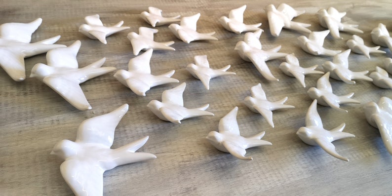 Op de afbeelding: Een verzameling witte keramische vogelbeeldjes, elk met uitgespreide vleugels, gerangschikt op een lichtgekleurd houten oppervlak. De vogels bevinden zich in verschillende houdingen, die vlucht suggereren, en zijn uniform wit met een glanzende afwerking.