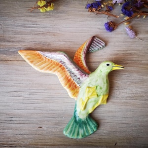 May include: A ceramic hummingbird sculpture with outstretched wings. The bird features a green body, yellow accents, and orange and white wings. The beak is yellow. The sculpture is set against a wooden background.