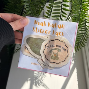 May include: A sticker pack with the text "Noah Kahan Sticker Pack." The stickers have a floral design and the text "I'll Call Your Mom" and "The Great". The stickers are in a clear plastic sleeve.